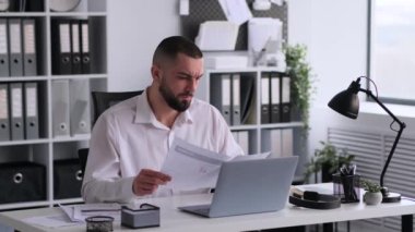 Genç beyaz erkek ofis çalışanı evrak işleriyle uğraşıyor, omuz silkiyor, modern ofiste dizüstü bilgisayarla oturuyor. Belgeler ve raporlar analiz ediliyor.