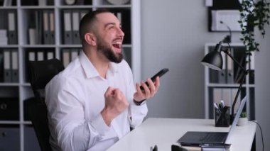 Şaşırmış, mutlu Kafkas ofis çalışanı telefonda konuşuyor ve ofiste çalışırken başarıyı ya da başarılı anlaşmayı kutluyor. Pozitif sonuç, finansal büyüme.