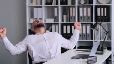 Kafkas erkek ofis çalışanı modern ofiste dizüstü bilgisayarla çalışırken başarılı sonuçları kutluyor. Neşeli zafer, zafer ya da zafer.