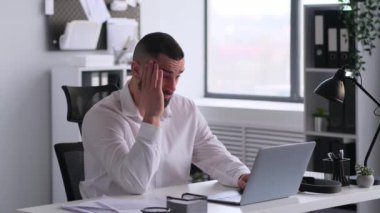 Masasında dizüstü bilgisayarla çalışan, ciddi bir beyaz erkek çalışanın baş ağrısı ya da migreni var. Yorgunluk, tükenme kavramı.