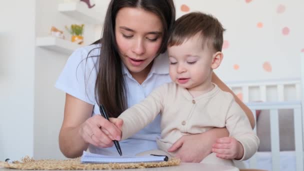 快乐的小儿子和笑着的白人年轻妈妈一起在儿童游戏室用钢笔和笔记本画画 母亲教育儿子 — 图库视频影像