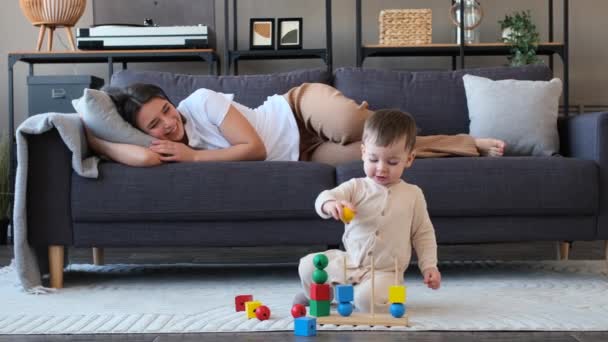 笑顔の若い白人母親はソファーでリラックスし リビングルームの床にカラフルなパズルのおもちゃで遊んでいる彼女の幼い息子を見て — ストック動画