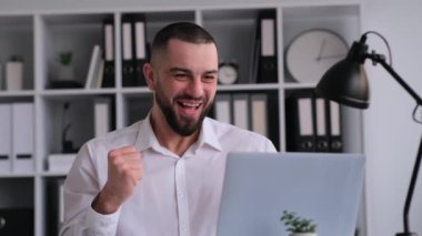 Mutlu ve şaşkın beyaz erkek, finansal büyümeyi, kazanmayı ya da başarıyı kutluyor, dizüstü bilgisayarla çalışıyor, ofisinde oturuyor..