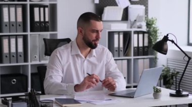 Beyaz erkek ofis çalışanı ya da yönetici, ofiste dizüstü bilgisayarla çalışır ve çevrimiçi konferans düzenler, evrak işleriyle uğraşır ve uzaktan iletişim kurar..