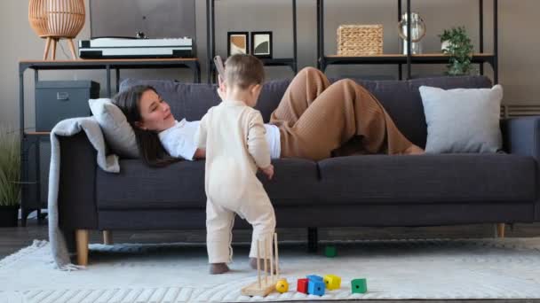 幸せな若いママはソファーに横たわり 携帯電話で休んで 彼女の幼い息子がおもちゃで遊んでリビングルームで笑っているのを見ます — ストック動画