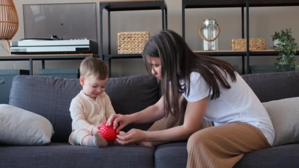 stock video Caucasian boy toddler and smiling mom or babysitter playing together with red ball and laughing on sofa in living room. Family playtime.