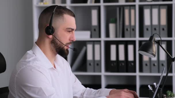 ハッピーコーカサス男性コールセンターオペレーターは ヘッドセットを着用し ラップトップを使用してオフィスでクライアントと話します テレマーケティングと通信コンセプト — ストック動画