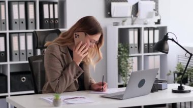 Beyaz kadın ofis çalışanı müşteriyle telefonda konuşuyor, dizüstü bilgisayar kullanıyor, evrak işleriyle uğraşıyor, modern ofiste masa başında oturuyor. Uzaktan danışman, profesyonel danışman.