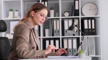 Belgeler ve dizüstü bilgisayarla çalışan, modern ofiste masa başında oturan, odaklanmış beyaz bir kadın. Planlama bütçesi, muhasebe işi, mali raporları denetleme.