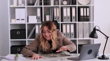 Gülümseyen beyaz kadın çalışan havaya para saçıyor, ofiste masa başında oturuyor. Zenginliği kutlamak, kumarda kazanmak, finansal başarı.