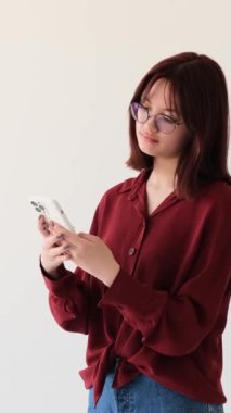 Şaşırmış ve mutlu beyaz kız beyaz arka planda cep telefonundan iyi haberler alıyor. İnternet taraması, online sosyal medya. Dikey video.