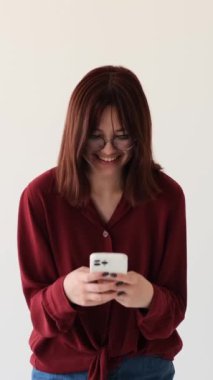 Gülümseyen ve mutlu, beyaz arka planda mesajlaşan ve gülen akıllı telefonlu beyaz kız. Sosyal medyada sohbet ediyorlar. Dikey video.