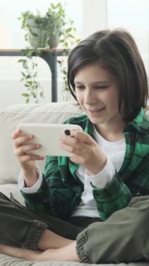 Evde cep telefonu kullanarak video oyunu oynayan mutlu beyaz okul çocuğu. Çevrimiçi, eğlence ve rekabet. Dikey video.