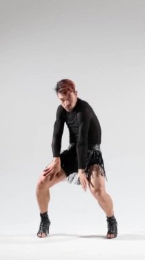 Enerjik ve yetenekli beyaz adam deri etek ve yüksek topuklu ayakkabı giyiyor, beyaz arka planda şehvetli dans ediyor. Dikey video.