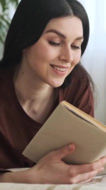 Mutlu, gülümseyen, beyaz bir kadının portresi yatakta dinleniyor ve kitap okuyor, hafta sonunu evde geçiriyor. Dikey video.