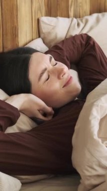 Kafkasyalı mutlu bir kadın yatakta uyanıyor ve evde esniyor. Sabah rutininin tadını çıkarıyor, rahat bir yatakta dinleniyor. Dikey video.