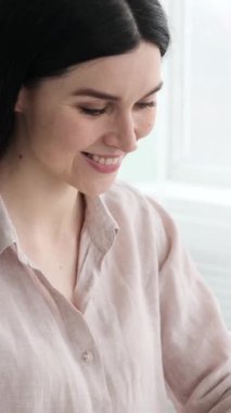 Ofiste çalışırken laptopta yazan, mesajlaşan ya da mesajlaşan gülümseyen Kafkasyalı bir kadın resmi. Dikey video.