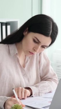 Yorgun ve ciddi bir iş kadını, baş ağrısı, evrak işleriyle meşgul, fazla mesai yapıyor. Yorgunluk ve tükenmişlik kavramı. Dikey video.