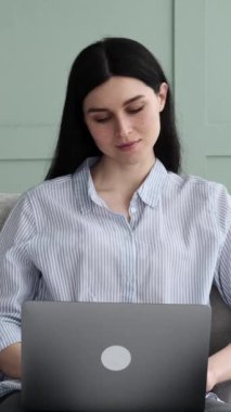 İnternette eğlenmek için dizüstü bilgisayar kullanan neşeli bir kadın. Oturma odasındaki kanepede oturuyor. Boş zaman konsepti sırasında uzaktan çalışma ya da rahatlama. Dikey video.