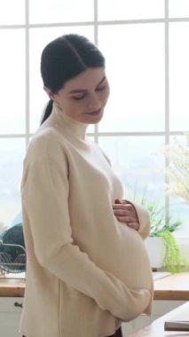 Neşeli Kafkasyalı hamile kadın ev mutfağında gülümseyerek karnını okşuyor. Bebek ve gelecekteki hamilelik konsepti bekliyor. Dikey video.