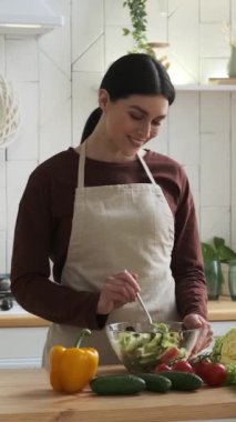Mutfakta sebze salatası pişiren arkadaş canlısı ve gülümseyen beyaz kadın. Kasede salata karıştırmak. Mutfak ve vejetaryen gıda konsepti. Dikey video.