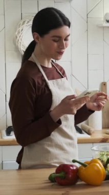 Neşeli beyaz kadın taze sebze salatası yiyor ve cep telefonunu ev mutfağında internet iletişimi ve eğlence için kullanıyor. Dikey video.