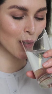 Camdan temiz su içen ve evde kameraya bakan mutlu bir beyaz kadının portresi. Su dengesini tazelemek ve kurtarmak. Dikey video.