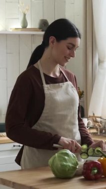 Önlük giyen mutlu ve pozitif beyaz kadın evde sebze salatası hazırlıyor. Sağlıklı yemek ve mutfak konsepti. Dikey video.