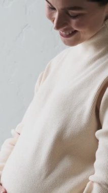 Evde karnını okşayan hamile, beyaz bir kadının portresini yakından çek. Gelecekteki ebeveynlik ve annelik, şefkat kavramı. Dikey video.