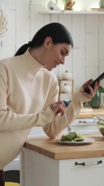 Neşeli hamile, beyaz kadın taze salata yiyor, akıllı telefon kullanıyor ve evde karnını okşuyor. Sosyal medya ve online eğlence. Dikey video.