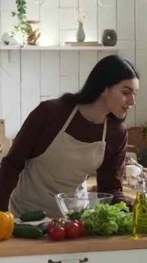 Önlük giymiş, yemek pişiren, salata için sebze kesen neşeli beyaz kadın. Sağlıklı yemek pişiren olumlu ev hanımı. Dikey video.