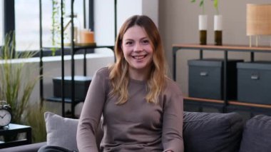 Neşeli ve aktif Kafkasyalı genç bir kadın baş parmağını kaldırıyor ve dans ediyor. Oturma odasındaki kanepede oturuyor. El işareti gibi..