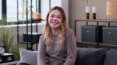 Tatmin olmuş beyaz bir kadın iki eliyle birlikte oturma odasındaki kanepede oturarak iyi niyet göstergesi gösteriyor. Memnuniyet ve tavsiye.