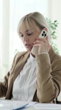 Yoğunlaşmış Kafkasyalı iş kadını modern ofiste evrak işleriyle uğraşıyor ve telefonla konuşuyor. Çevrimiçi sohbet ve sohbet. Dikey video.