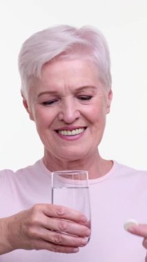 Yaşlı heyecanlı beyaz kadın köpüklü, köpüren bir tableti beyaz arka planda suyla birlikte bardağa atıyor. İlaç ve sağlık sigortası konsepti. Dikey video.