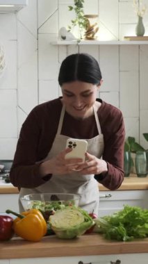 Gülümseyen beyaz kadın vlogger, blogcu sosyal medya için taze ve sağlıklı salatayı fotoğraflıyor mutfakta cep telefonu kullanıyor. Dikey video.