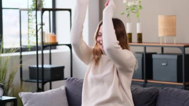 Neşeli ve gülümseyen beyaz bir kadın kanepede dinleniyor müzik dinliyor ve evde dans ediyor. Eğlence ve kutlamayla meşgul, boş zamanlarını harcıyor..