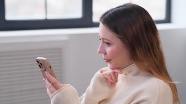 Dost canlısı ve neşeli Kafkasyalı kadın internette sohbet ediyor, internette geziniyor ve gülüyor, evdeki oturma odasında telefonla kanepede dinleniyor..