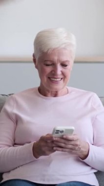 Gülümseyen ve iyimser Kafkas yetişkin kadın portresi evde telefonla kameraya bakıyor. İnternete göz atıyorum. Dikey video.