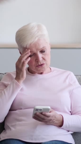 在网上用电话阅读坏消息 在家里客厅里患有偏头痛的悲伤而不健康的高加索老年妇女 垂直录像 — 图库视频影像