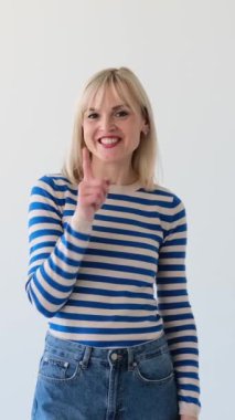 Beyaz arka planda dedikodu veya sır tutmak için sessiz bir jestle flört eden mutlu beyaz bir kadının olumlu portresi. Dikey video.