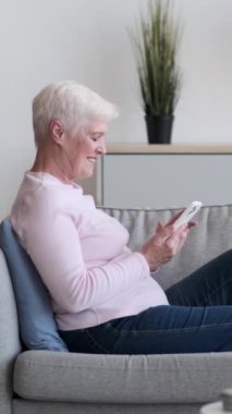 Telefon kullanan, sosyal medyada mesajlaşan ya da internette gezinen, evdeki kanepede uzanan arkadaş canlısı, Kafkasyalı yaşlı kadın. Boş zaman gevşemesi. Dikey video.
