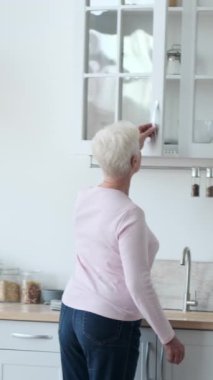 Ev mutfağında yemek masası kuran yaşlı mutlu beyaz kadın. Sofra takımı ile servis yapmak ve ziyafet hazırlamak. Dikey video.