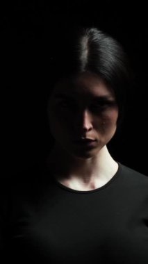 Modaya uygun ve melankolik bir kadın, kontrastlı ışıkta, siyah arka plandan kameraya bakıyor. Dramatik, düşünceli kadın portresi. Dikey video.