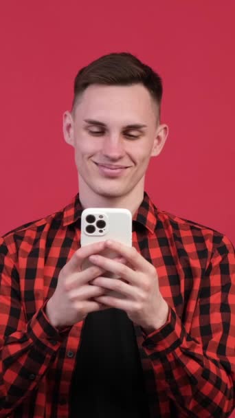 広い笑顔で陽気な白人男性のポジティブな肖像画 携帯電話のメッセージを保持したり 赤い背景でオンラインで閲覧 バーティカルビデオ — ストック動画