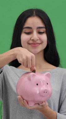 Heyecanlı Hintli kadın yeşil arka planda kumbaraya para koyuyor. Para tasarrufu ve finansal başarı kavramı. Dikey video.