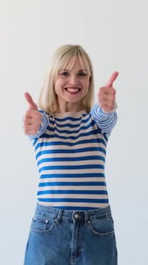 Beyaz arka planda mutlu ve tatmin olmuş beyaz bir kadın baş parmağını gösteriyor ya da jest yapıyor ve dans ediyor. Doğrulama ve anlaşma konsepti. Dikey video.