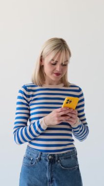 Sosyal medyada akıllı telefonuyla mesajlaşan ve beyaz arka planda kameraya gülümseyen arkadaş canlısı ve kaygısız beyaz kadın. Dikey video.