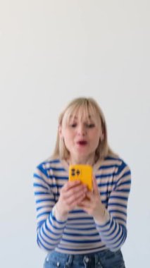 Şaşırmış Kafkasyalı kadın, boş zamanlarını telefon kullanarak geçiriyor, internette iyi haberler alıyor ve şaşırıyor, beyaz arka planda elde edilen başarıların tadını çıkarıyor. Dikey video.