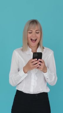 Neşeli kadın ofis çalışanı çevrimiçi mesajlaşıyor ve gülüyor, mavi arka planda cep telefonuyla vakit geçiriyor. Dikey video.